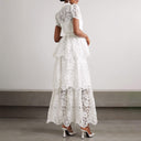 Back view of a white tiered lace maxi dress by self-portrait. The dress features a sheer lace high neck and high back with short sleeves with an opaque fitted bust. The bottom of the dress is divided into 3 tiers of lace overlapping each other.